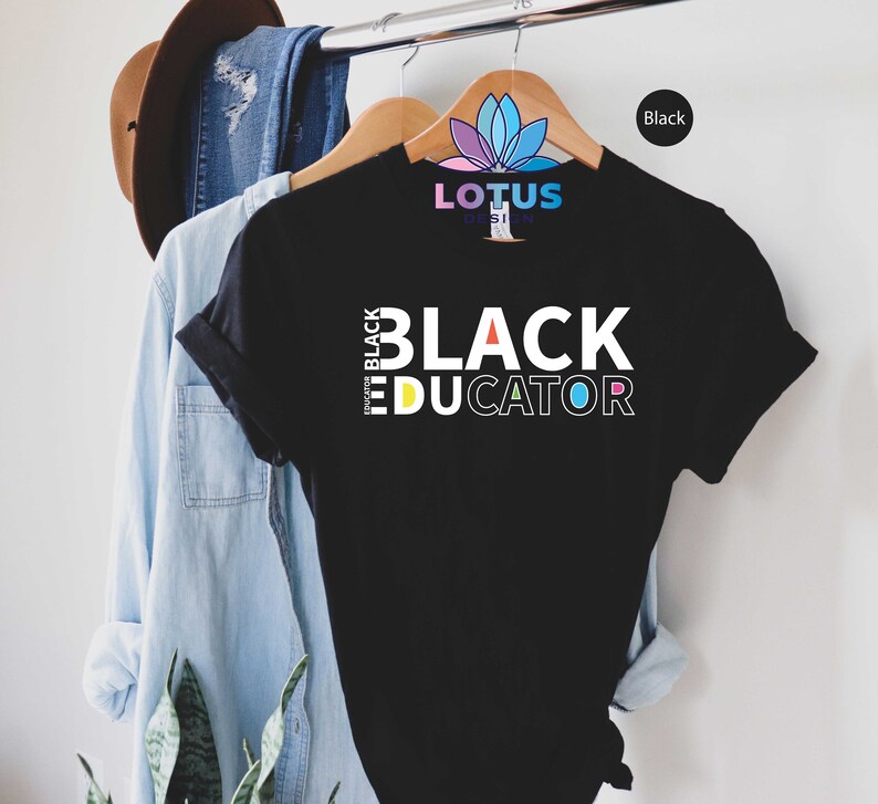 May include: Black t-shirt with the text "I'm Black Educator" in white letters with a rainbow color scheme. The shirt is hanging on a wooden hanger.