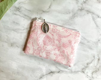 Pode incluir: Uma pequena bolsa com estampa floral rosa com zíper prateado e pingente prateado. A bolsa é retangular e apresenta um delicado design floral repetido em tons de rosa sobre um fundo branco. O pingente tem formato oval.