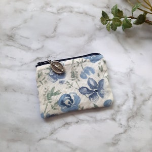 May include: A small zippered pouch with a floral pattern. The pouch is white with blue flowers and green leaves. A silver charm with a religious image is attached to the zipper.