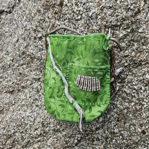 May include: A small green fabric pouch featuring a leaf pattern, stitched with light green thread. The pouch is adorned with a curved piece of driftwood, a small pocket, and a textured, striped shell fragment attached to the front surface.
