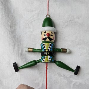 May include: A wooden Christmas ornament in the shape of a soldier. The ornament has a green hat, green body, and black boots. The soldier's face has a mustache and a red nose. The arms and legs are jointed and move freely. The ornament hangs from a red string.