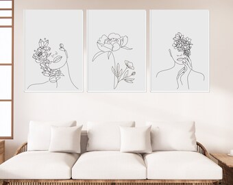 May include: Three black and white line art prints featuring a woman's face with flowers, a single flower, and a woman's face with flowers. The prints are framed and hung on a white wall above a white couch.
