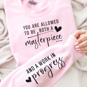May include: Pink sweatshirt with the text "You are allowed to be both a masterpiece and a work in progress" in black lettering with a small heart graphic.