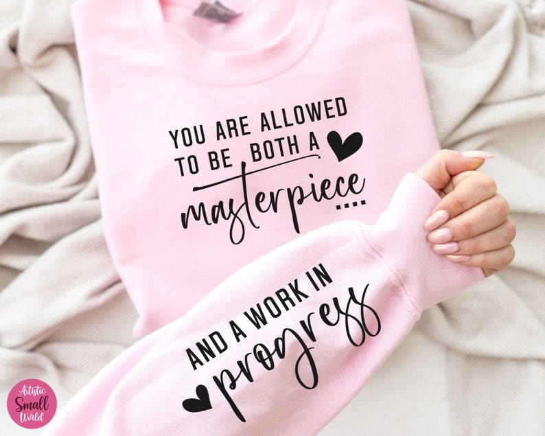 May include: Pink sweatshirt with the text "You are allowed to be both a masterpiece and a work in progress" in black lettering with a small heart graphic.