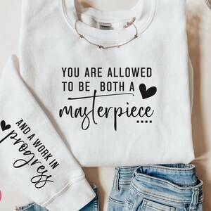 May include: White sweatshirt with the text "YOU ARE ALLOWED TO BE BOTH A masterpiece AND A WORK IN progress" printed on it. The text is in black and there are small hearts on either side of the text.