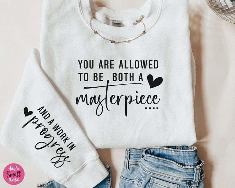 May include: White sweatshirt with the text "YOU ARE ALLOWED TO BE BOTH A masterpiece AND A WORK IN progress" printed on it. The text is in black and there are small hearts on either side of the text.