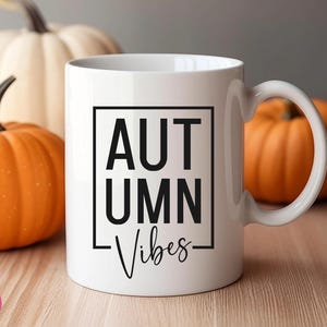 Puede incluir: Taza de cer&aacute;mica blanca con un gr&aacute;fico negro que dice "Autumn Vibes" en un cuadro. La taza est&aacute; sobre una superficie de madera con dos calabazas naranjas.