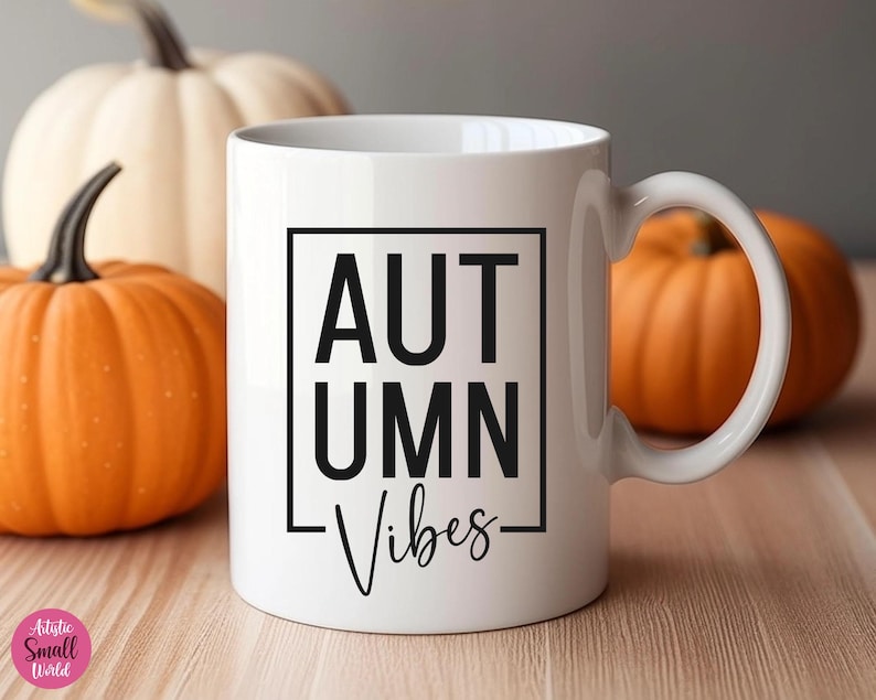 Puede incluir: Taza de cer&aacute;mica blanca con un gr&aacute;fico negro que dice "Autumn Vibes" en un cuadro. La taza est&aacute; sobre una superficie de madera con dos calabazas naranjas.