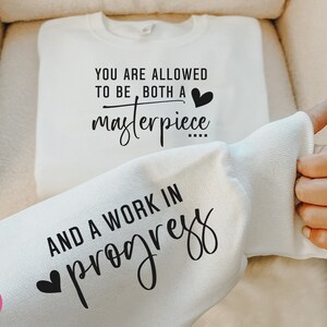 May include: White sweatshirt with a black graphic print that reads "You are allowed to be both a masterpiece and a work in progress" with a small heart icon above the word "masterpiece" and below the word "work".