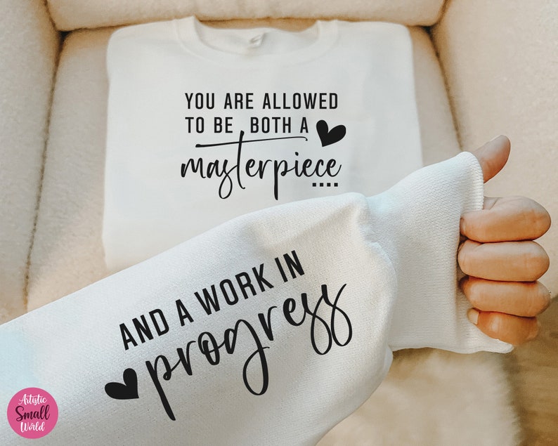 May include: White sweatshirt with a black graphic print that reads "You are allowed to be both a masterpiece and a work in progress" with a small heart icon above the word "masterpiece" and below the word "work".