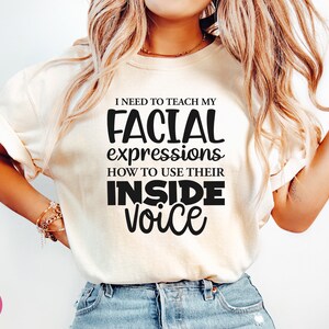 May include: A white t-shirt with black text that reads "I need to teach my facial expressions how to use their inside voice."