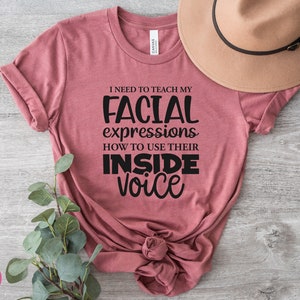 May include: A pink t-shirt with the text "I need to teach my facial expressions how to use their inside voice" printed in black.