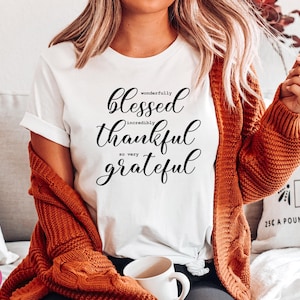 Puede incluir: Camiseta blanca con texto negro que dice "wonderfully blessed incredibly thankful so very grateful". La camiseta la lleva puesta una persona con el pelo largo y rubio y un c&aacute;rdigan marr&oacute;n.