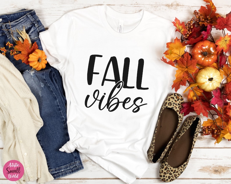 Puede incluir: Camiseta blanca con el texto "Fall vibes" en letras negras. La camiseta se muestra sobre una superficie de madera blanca con decoraciones de oto&ntilde;o, incluyendo jeans, una chaqueta de punto y una guirnalda de hojas de oto&ntilde;o y calabazas.