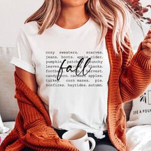 以下が含まれることがあります： 「fall」という単語が黒色の書体で書かれた白いTシャツ。その周りを、「心地よいセーター」、「スカーフ」、「ジーンズ」、「ブーツ」、「アップルサイダー」、「パンプキンパッチ」、「カリカリの葉」、「ありがとう」、「フットボール」、「収穫」、「スイートコーン」、「キャラメルアップル」、「ターキー」、「コーンメイズ」、「パイ」、「焚き火」、「干し草乗り」、「秋」などの秋のテーマの単語が囲んでいます。