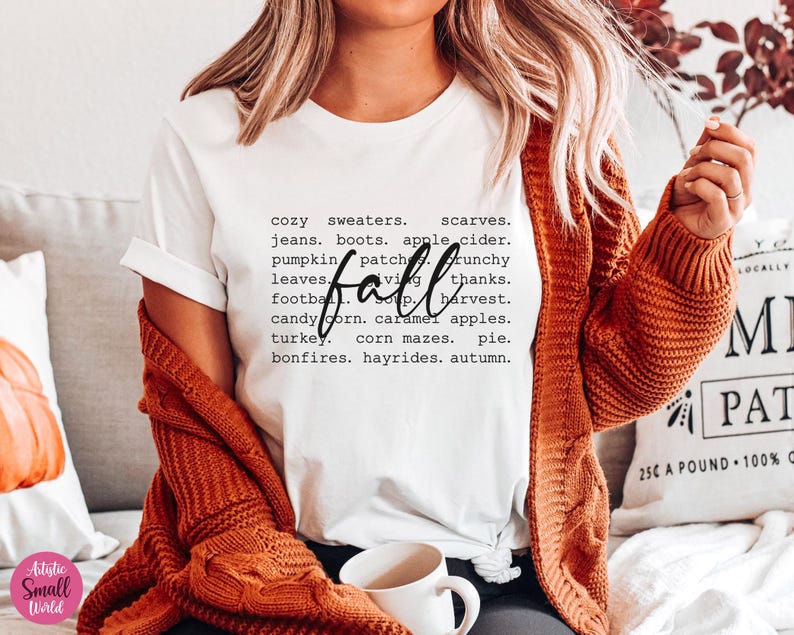 以下が含まれることがあります： 「fall」という単語が黒色の書体で書かれた白いTシャツ。その周りを、「心地よいセーター」、「スカーフ」、「ジーンズ」、「ブーツ」、「アップルサイダー」、「パンプキンパッチ」、「カリカリの葉」、「ありがとう」、「フットボール」、「収穫」、「スイートコーン」、「キャラメルアップル」、「ターキー」、「コーンメイズ」、「パイ」、「焚き火」、「干し草乗り」、「秋」などの秋のテーマの単語が囲んでいます。