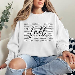 以下が含まれることがあります： 白いスウェットシャツに、大きな黒い文字で「fall」と書かれており、その周りを秋の言葉を並べたリストが囲んでいます。言葉には、居心地の良いセーター、スカーフ、ジーンズ、ブーツ、アップルサイダー、パンプキンパッチ、サクサクの葉、フットボール、感謝祭、収穫、キャンディーコーン、キャラメルアップル、ターキー、コーンメイズ、パイ、焚き火、馬車に乗る、秋などが含まれています。