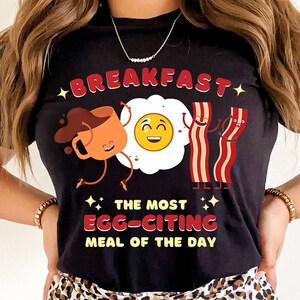 Puede incluir: Camiseta negra con una ilustraci&oacute;n de dibujos animados de una taza de caf&eacute;, un huevo frito y tocino. El texto dice "Breakfast, The Most Egg-citing Meal of the Day".