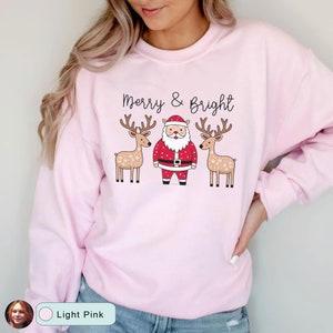 May include: Light pink sweatshirt with the text "Merry & Bright" above a Santa Claus and two reindeer. The Santa Claus is wearing a red suit with a black belt. The reindeer have antlers and snowflake patterns.