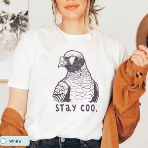 May include: White t-shirt featuring a black and white illustration of a pigeon wearing sunglasses. The text "stay coo." is printed below the bird. The model is wearing a brown jacket.
