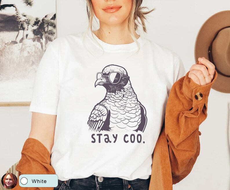 May include: White t-shirt featuring a black and white illustration of a pigeon wearing sunglasses. The text "stay coo." is printed below the bird. The model is wearing a brown jacket.