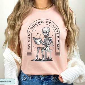 May include: A pink t-shirt with a graphic of a skeleton reading a book. The graphic is surrounded by an arch with the text "So Many Books, So Little Time".