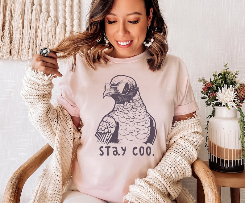 May include: A light pink t-shirt featuring a graphic of a bird wearing sunglasses and the text "STAY COO." The shirt has short sleeves and a crew neck. The shirt is made of a soft material.