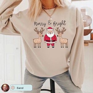 May include: Sand-colored sweatshirt with the text "Merry & Bright" above a Santa Claus graphic flanked by two reindeer. The Santa Claus is wearing a red suit and hat. The reindeer have brown antlers and are decorated with white snowflakes.