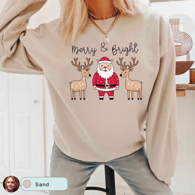 May include: Sand-colored sweatshirt with the text "Merry & Bright" above a Santa Claus graphic flanked by two reindeer. The Santa Claus is wearing a red suit and hat. The reindeer have brown antlers and are decorated with white snowflakes.