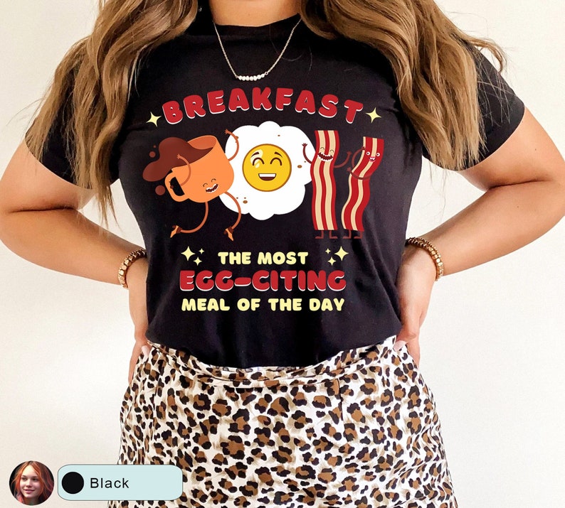 Puede incluir: Camiseta negra con una ilustraci&oacute;n de dibujos animados de una taza de caf&eacute;, un huevo frito y tocino. El texto dice "Breakfast, The Most Egg-citing Meal of the Day".