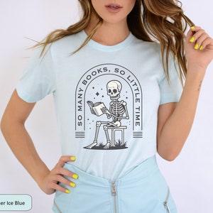 May include: A light blue t-shirt with a graphic of a skeleton reading a book. The graphic is surrounded by a circle with the text "So many books, so little time".