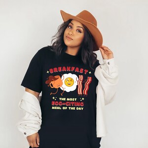 Puede incluir: Camiseta negra con una ilustraci&oacute;n de dibujos animados de una taza de caf&eacute;, un huevo y tocino. El texto dice "Breakfast, The Most Egg-citing Meal of the Day".