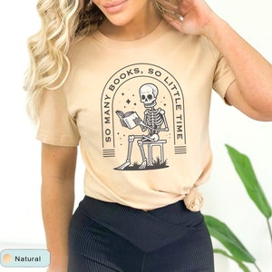 May include: A tan t-shirt with a black and white graphic of a skeleton reading a book. The graphic says "So many books, so little time".
