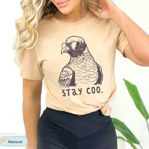 May include: A tan t-shirt featuring a cartoon pigeon wearing sunglasses. The pigeon is drawn in black and white, with the words "STAY COO." printed below. The shirt is a neutral color and has short sleeves.