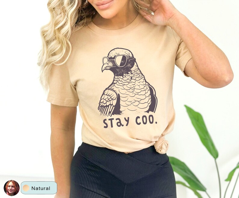 May include: A tan t-shirt featuring a cartoon pigeon wearing sunglasses. The pigeon is drawn in black and white, with the words "STAY COO." printed below. The shirt is a neutral color and has short sleeves.