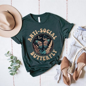 May include: A dark green t-shirt with the text "Anti-Social Butterfly" in a retro font. The text is surrounded by a circle with a butterfly and floral design.