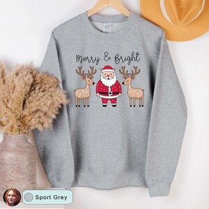 May include: Sport grey sweatshirt with the text "Merry & Bright" above a graphic of Santa Claus and two reindeer. Santa is wearing a red suit and hat with a black belt. The reindeer have brown antlers and snowflake patterns.