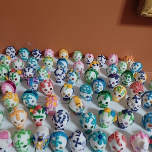 May include: A collection of colorful, decorated sugar skulls, each with unique designs. The skulls feature various colors like blue, green, pink, and yellow, with intricate icing details and some with small embellishments. They are arranged on a white surface.