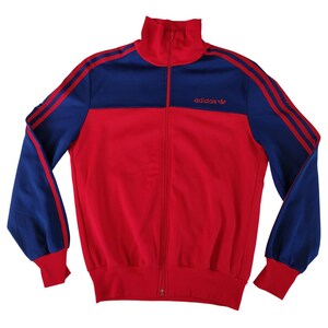 Peut inclure: Veste de survêtement Adidas rouge et bleu avec fermeture éclair intégrale et col montant. La veste présente des manches bleues avec des rayures rouges et le logo Adidas sur la poitrine. Style vintage.