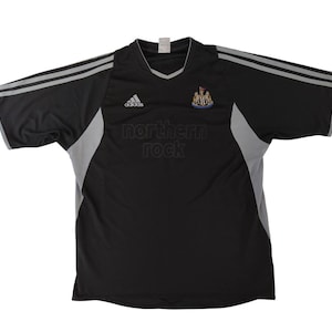 May include: Black and grey football shirt featuring the Adidas logo and the text "northern rock". The shirt has a V-neck collar and grey stripes on the sleeves. The Newcastle United crest is on the chest.