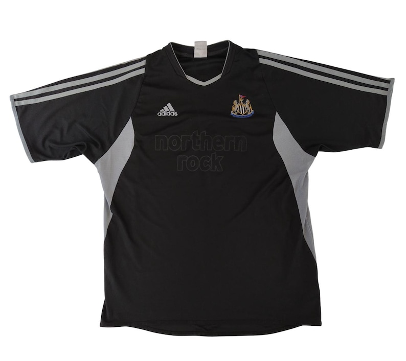 May include: Black and grey football shirt featuring the Adidas logo and the text "northern rock". The shirt has a V-neck collar and grey stripes on the sleeves. The Newcastle United crest is on the chest.