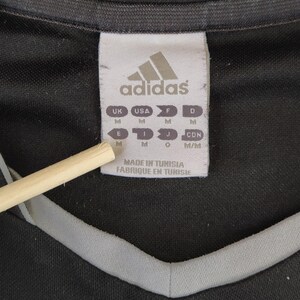 May include: A close-up shot of a black sports shirt featuring a grey neckline trim. The Adidas logo is displayed on a white label, alongside size markings for different regions. The label also states the shirt was made in Tunisia.