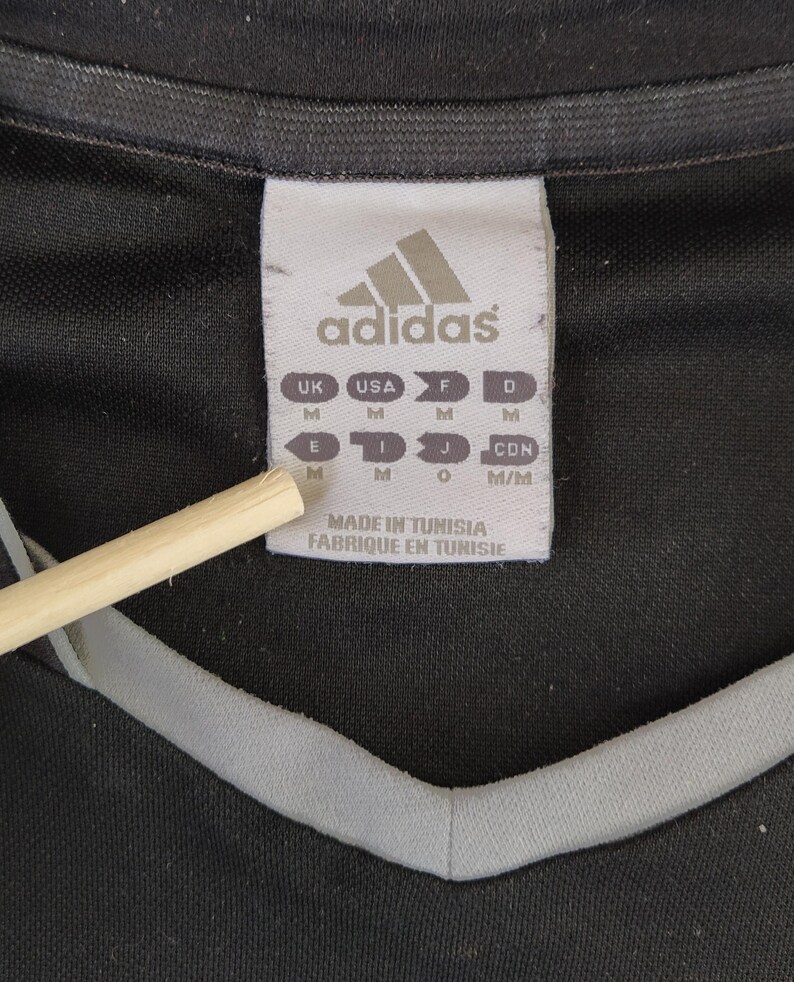 May include: A close-up shot of a black sports shirt featuring a grey neckline trim. The Adidas logo is displayed on a white label, alongside size markings for different regions. The label also states the shirt was made in Tunisia.