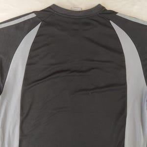 May include: A black athletic top featuring grey mesh side panels and grey stripes across the shoulders. The crew neck shirt is made from a lightweight, breathable fabric, ideal for sports and exercise activities.