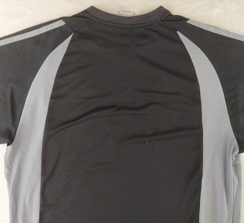 May include: A black athletic top featuring grey mesh side panels and grey stripes across the shoulders. The crew neck shirt is made from a lightweight, breathable fabric, ideal for sports and exercise activities.