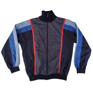 Op de afbeelding: Een marineblauwe trainingsjas met een volledige ritssluiting, hoge kraag en twee zijzakken. De jas heeft blauwe en lichtblauwe strepen op de mouwen en rode strepen aan de zijkanten. Het Adidas-logo is zichtbaar.