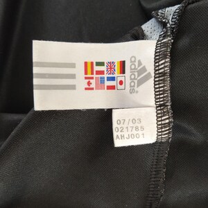 May include: A close-up of a black Adidas sports garment. A white label shows the Adidas logo, three stripes, and flags of different nations. The label also has the numbers 07/03, 021785, and AHJ001.
