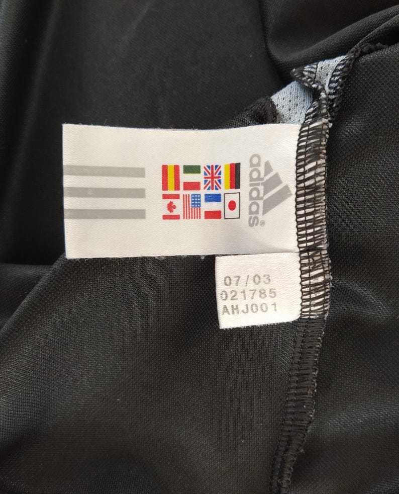 May include: A close-up of a black Adidas sports garment. A white label shows the Adidas logo, three stripes, and flags of different nations. The label also has the numbers 07/03, 021785, and AHJ001.