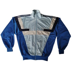 Op de afbeelding: Een vintage trainingsjack in blauw, lichtblauw en beige. De jas heeft een hoge kraag, een ritssluiting over de hele lengte en geribde manchetten en zoom. Het ontwerp omvat geometrische kleurgeblokte panelen op de voorkant en schouders.