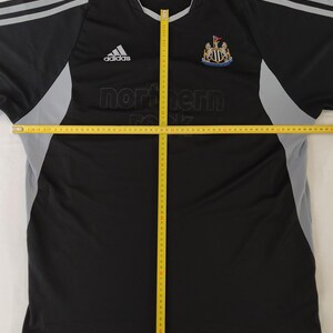 May include: Black and grey Adidas Newcastle United football shirt. The shirt displays the Adidas logo, the Newcastle United crest, and the text "northern rock". It has grey stripes on the shoulders.